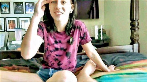Remi Jones smiling in purple tie-dye tee and denim shorts on bed
