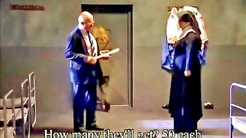 Bald older man in suit holds papers talking to veiled woman in dark robe in gray institutional room