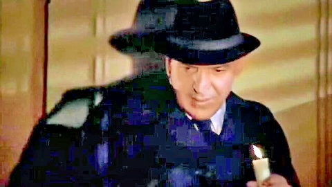 Mature man in dark suit and hat holds lit candle in shadowy BDSM scene