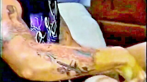 Tattooed muscular arm gripping white sheet in dimly lit retro bedroom scene