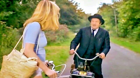 Vintage blonde woman in blue dress chats with older suited man on bicycle in rural countryside