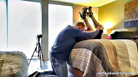 Husband kneels holding wife's legs high in black heels on bedroom bed ready for sex
