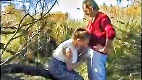 Blond twink kneels sucking older man's exposed cock outdoors in grassy field