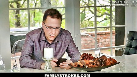 Mature man in glasses and checkered shirt intently texts on phone at sunroom table with candle and potpourri