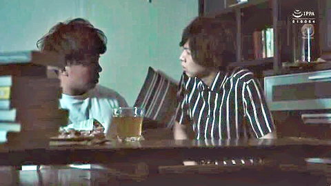 Two shaggy-haired Asian guys in casual shirts chat over pizza and beer at a book-filled table