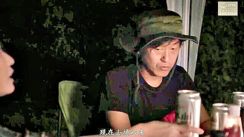 Japanese man in camo hat smiling holds beer cans at outdoor camping table with woman glancing
