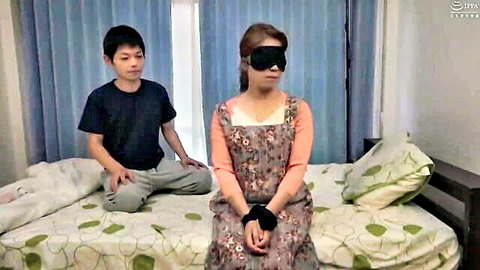 Blindfolded Asian stepmom with bound hands in floral dress awaits stepson's punishment