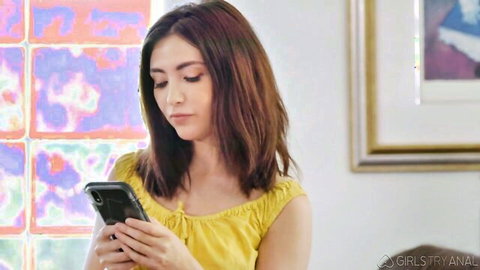 Gia Derza in yellow top holding phone in teasing threesome setup