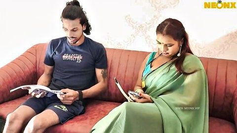 Sexy Indian MILF in sheer green saree sits with hunk in shorts on couch holding phone