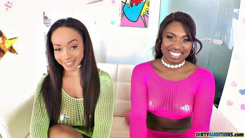 Two smiling ebony brunettes in sheer green and pink mesh tops teasing on casting couch