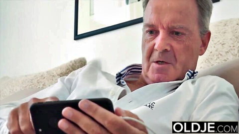 Older man in white shirt holding smartphone on couch, mouth open