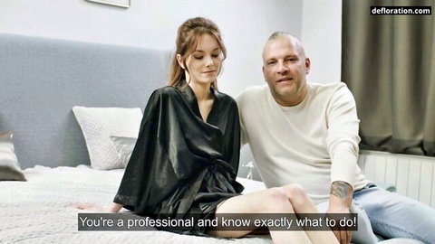 Skinny young brunette with small tits in open black robe sits smiling with older bald man on bed