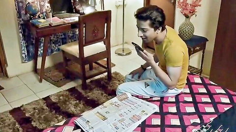 Indian man in yellow shirt sits on bed holding phone in cozy bedroom