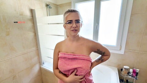 Blonde tattooed babe in glasses wrapped in pink towel teasing in bathroom
