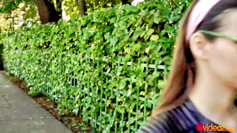 Sexy teen brunette in pink headband and green shades puckers lips teasingly by green hedge fence