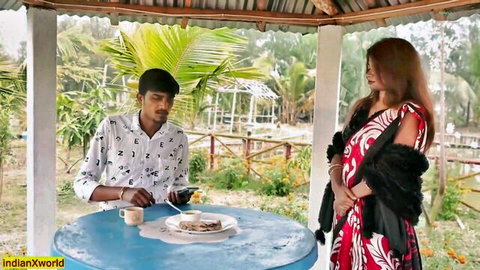 South Indian couple in steamy outdoor pavilion date, wife in red dress stands over seated husband at table