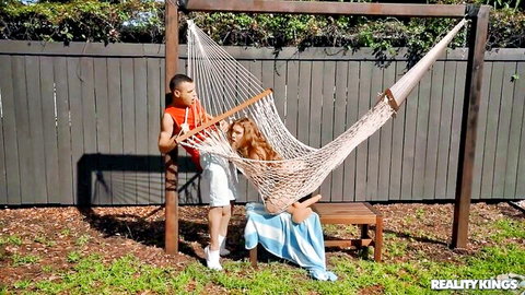 Redhead with small tits spreads shaved pussy on outdoor bench near hammock and muscular guy in shorts