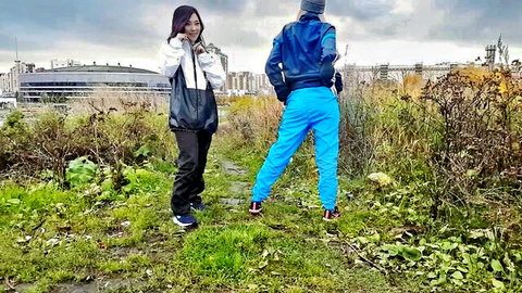 Asian brunette woman in shiny black pants smiles at camera with boyfriend in blue tracksuit outdoors