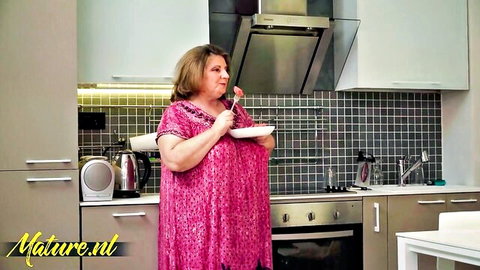 Chubby mature brunette MILF eating soup in pink dress in kitchen