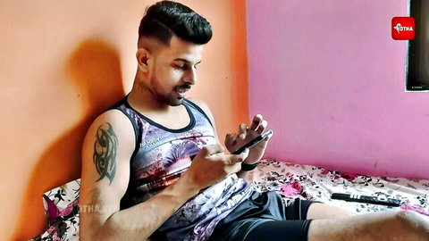 Tattooed Indian guy in tank top and shorts browsing phone on bed