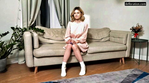 Blonde girl in lacy pink dress sits legs crossed on couch teasing