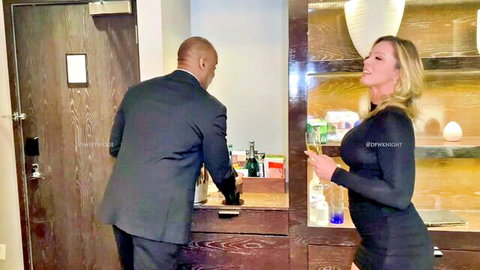 Blonde bombshell in tight black dress flirts with suited black bull holding champagne in hotel room