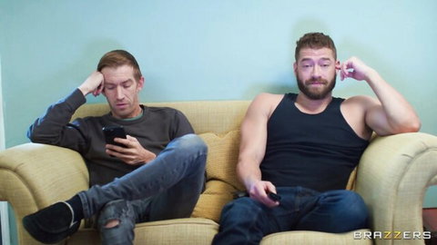 Two clothed studs sit on couch, redhead stressed on phone, bearded hunk with controller in tank top