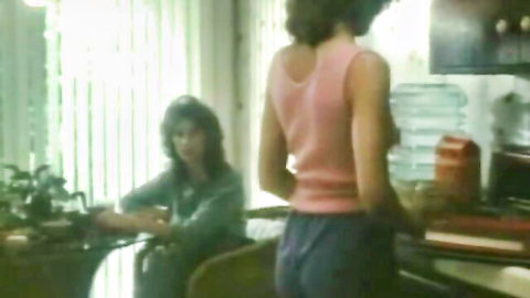 Vintage retro MILF in pink tank top stands teasingly over seated brunette woman in kitchen