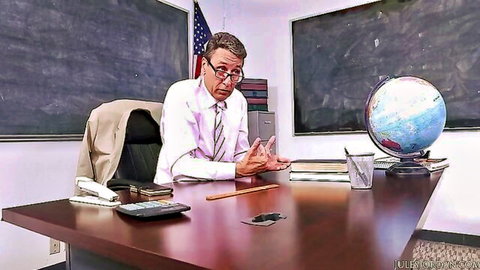 Clothed male teacher gesturing at desk in classroom with globe and flag