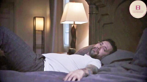 Bearded tattooed man lying asleep on bed with gray pants pulled down to thighs