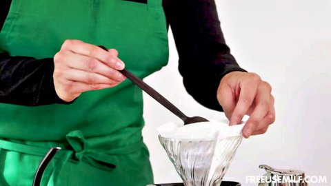 Barista in green apron stirring thick white foam into glass pour-over coffee dripper