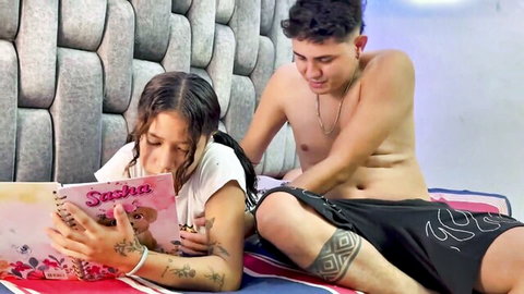 Tattooed tanned Latina teen reads pink Sasha notebook while shirtless guy embraces her from behind in bed