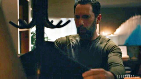 Bearded man in green shirt standing by coat rack in dimly lit room