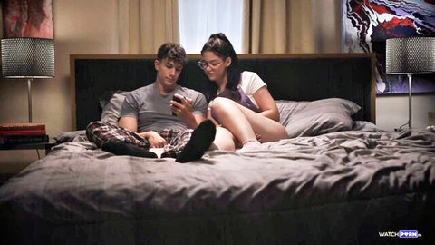 Stepsister in white tee and socks sits legs spread with stepbrother on bed as he checks phone