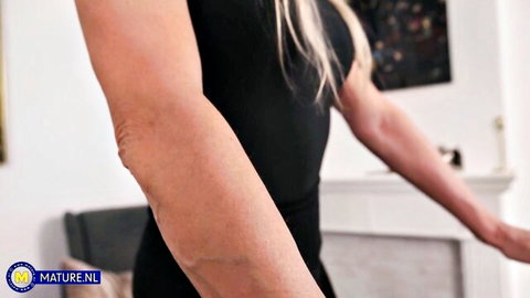 Seductive blonde MILF stretches toned arms in tight black dress by fireplace