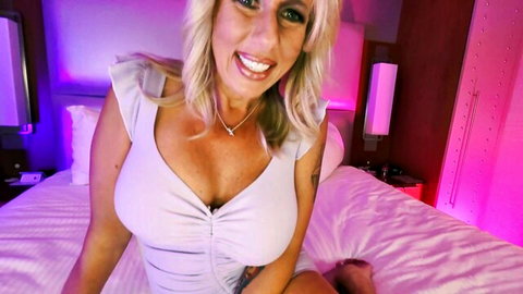 Curvy blonde MILF with huge tits smiling seductively in low-cut white top on bed