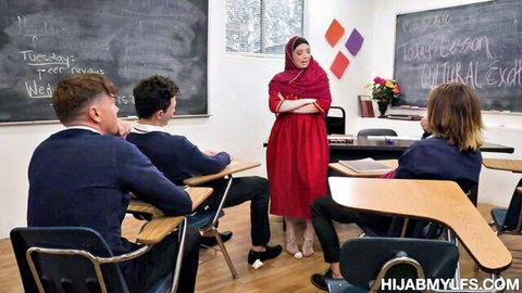 Hijab teacher in red dress stands arms crossed before uniformed students in classroom
