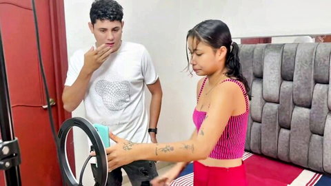 Tattooed Latina teen in pink crop top and red shorts holds ring light with surprised boyfriend in bedroom