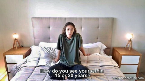 Luna from Argentina Casting sitting casually on bed in green top and jeans