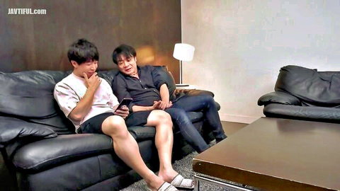 Two young East Asian guys lounging on black leather couch holding phones teasingly