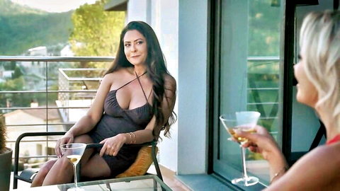 Busty brunette MILF in low-cut dress and blonde sipping martinis on balcony showing deep cleavage