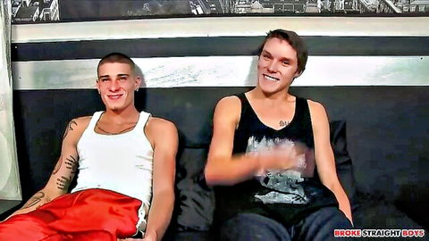 Two smiling young gay guys in tank tops and shorts sitting on couch teasing for action