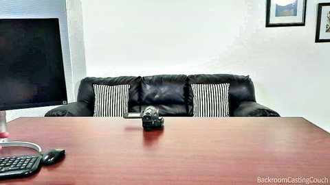 Empty Backroom Casting Couch setup with black leather sofa, wooden desk, camera, and monitor ready for amateur audition.