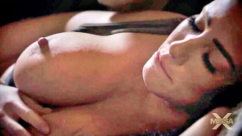 Busty brunette lying back with hard cock head on her exposed tit and nipple, eyes closed smiling in ecstasy