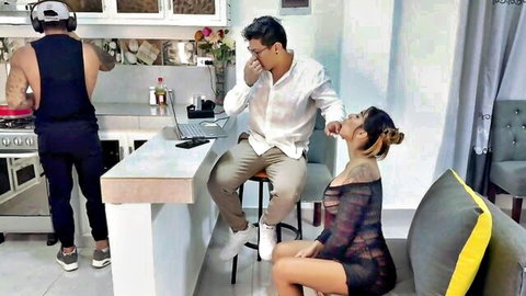 Blonde girlfriend in sheer black dress teases boyfriend on lap while muscular husband cooks in kitchen cuckold style
