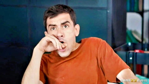 Shocked man in orange shirt leans on hand with open mouth in Brazzers scene
