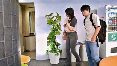 Young skinny Japanese brunette with long hair and big tits stands by plant as guy approaches her in lounge