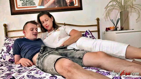 Mature brunette MILF smiles seductively cuddling young stud on purple bed