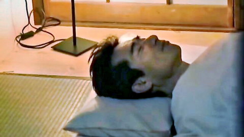 Shirtless Asian man lying asleep on futon in dimly lit Japanese room
