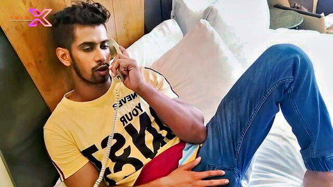 Handsome South Asian guy lounging on hotel bed talking on phone in yellow tee and jeans